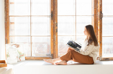 Young woman sits on a sunny windowsill in front of huge window, barefoot and relaxed, looking into the camera with a soft smile. Natural light illuminates the scene, warm and serene atmosphere