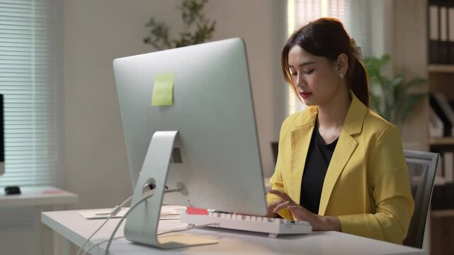 Asian office worker in a yellow jacket types on a keyboard while focused on tasks in a modern office environment