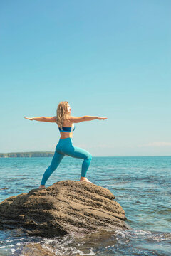 Woman practicing yoga in vira bhadrasana two warrior pose at cliff beach blue sea sky