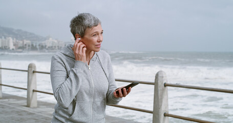 Mature woman, phone and earphones on promenade with music, reading and workout in morning. Person, smartphone and listening on mobile app with running, streaming and subscription by ocean in Italy