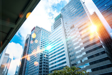 A low angle view of tall buildings in a city, the sun shining through the glass.