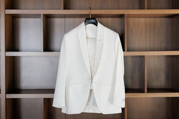 The bride's elegant jacket hangs on a hanger in the room. A white women's jacket hangs on a hanger on a background of wooden shelves. White women's suit jacket