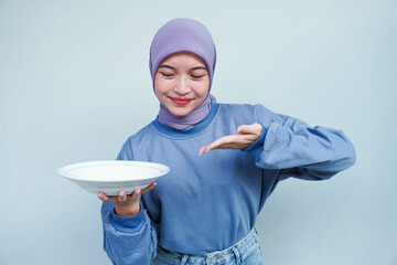 A smiling Asian Muslim woman is fasting and hungry and holding and pointing to a plate.