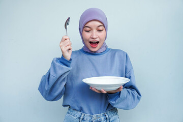 A smiling Asian Muslim woman is fasting and hungry and holding and pointing to a plate.