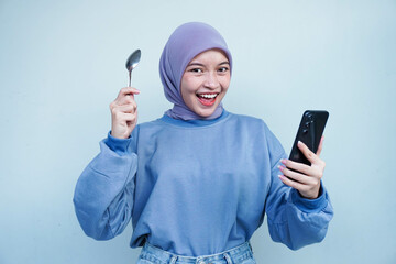 Happiness Asian Muslim woman is fasting and hungry and holding cutlery while looking phone get idea about what to eat. Online food concept.