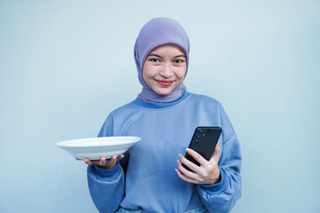 Happiness Asian Muslim woman is fasting and hungry and holding cutlery while looking phone get idea about what to eat. Online food concept.