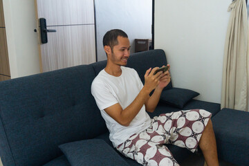 Obraz premium Young asian men wearing white shitr is holding smart phone with smile on sofa.