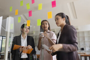 Asian businesswoman teamwork planning with sticky notes and new ideas on a glass wall, Business people brainstorming strategy and new ideas concept, startup worker meeting,