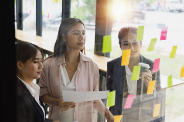 Asian businesswoman teamwork planning with sticky notes and new ideas on a glass wall, Business people brainstorming strategy and new ideas concept, startup worker meeting,