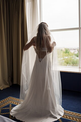 The bride puts on her dress while standing by the window.