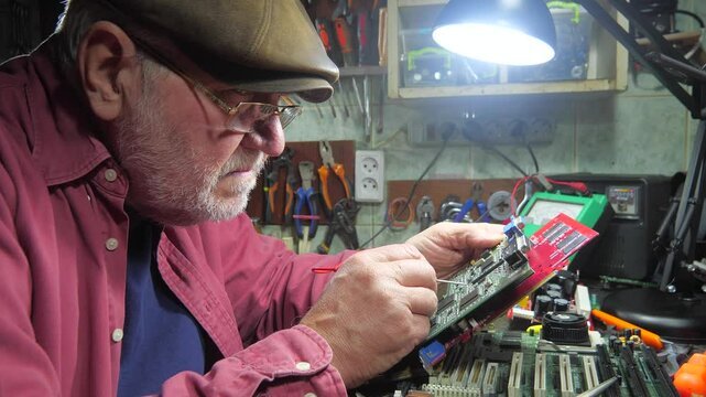 An old master repairs computer components in a workshop. Using a soldering iron and other tools, he repairs and adjusts broken electronics.