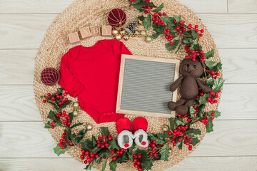 Festive baby announcement setup featuring a baby onesie, a letterboard, and seasonal holiday decorations on a soft background. Ideal for Christmas-themed baby reveal or holiday family announcement.