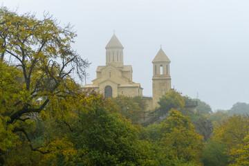 Naklejka premium Church of St. Ekvtime and bell tower in Kiketi village on a foggy morning. Trees with autumn leaves, bushes in the foreground