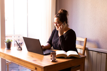 Young, white, smiling woman working from home in her private office. With a computer, a book, pens and a cell phone. Wooden table, black clothes.