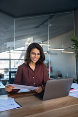 Smiling latin hispanic young business woman working on laptop computer reading financial document...