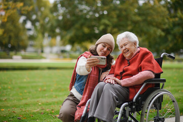 Granddaughter on an autumn walk in the park with her grandmother in wheelchair, taking selfie with...