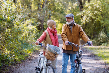 Obraz premium Lovely elderly couple on a walk in the forest, pushing their bikes side by side. Seniors in love on stroll through autumn nature, enjoying a peaceful moment together.