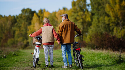 Rear view of elderly couple on a walk in the forest, pushing their bikes side by side. Seniors in love on stroll through autumn nature, enjoying a peaceful moment together.