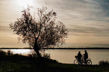 Lovely elderly couple on standing by lake with their bikes and looking at sunset, enjoying a...
