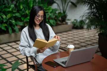 An attractive, calm Asian woman sits at an outdoor table at a coffee shop, focused on reading.