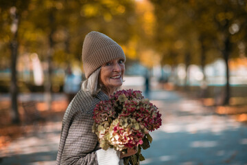 Portrait of a beautiful senior woman standing outdoors with bouquet of dried flowers. Autumn park...