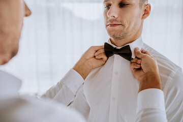 A man adjusts another man’s bow tie, focusing on detail. The close-up captures a moment of preparation, with both dressed in formal white shirts..