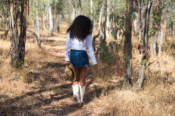Young, beautiful, Latin, brunette woman with curly hair wearing a white shirt, denim skirt and boots, holding a hat in one hand and touching herbs with the other hand. Photo taken from behind.