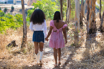 Two women, mother and daughter of Latin origin and brunettes walk hand in hand along a path between trees. Photo taken from behind. Mother's day concept.