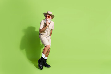 Fototapeta premium Elderly man in safari-style outfit poses against green backdrop in casual shorts and summer glasses.