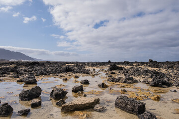 White sand beach with black rocks of volcanic origin. Mountains in the background and sky with big white clouds. Caletón Blanco, Lanzarote, Canary Islands, Spain