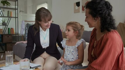 Medium long shot of cheerful worker of social aid service talking to little girl sitting next to mother at home