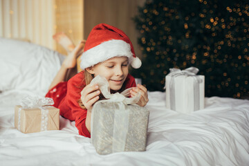 Cute 7-years old girl wearing Santa hat and holiday pajamas enjoying her a Christmas presents in a festive bedroom.