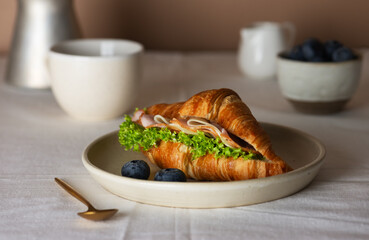 Croissant sandwich with ham, cheese and lettuce, with cup of coffee and blueberries on a table. Morning breakfast.