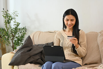 Satisfied young asian woman making a purchase online with credit card and digital tablet at home