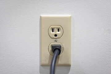 Wall outlet on a white wall with one plug connected. North American outlet with beige outlet plate