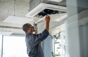 High up, fixing the ventilation. Repairman is indoors in the modern office