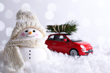 Cute decorative snowman and toy car with fir tree branches on artificial snow against light...