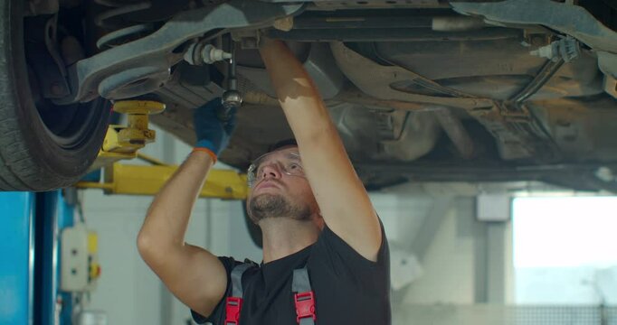 Car mechanic is working under a vehicle on a lift in service. Repairman is using ratchet. Specialist is wearing safety glasses.