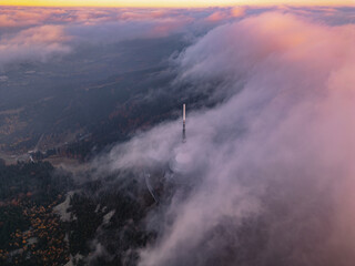 Fototapeta premium Dramatic clouds envelop the scenery during a vibrant sunset in Liberec, Czechia, highlighting the tower rising above the misty landscape, creating an enchanting atmosphere.