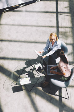 Two diverse business women sit in a large, bright office space and talk together and having a casual meeting. One of the women is taking notes on her laptop. On the table is paper with graphs. 