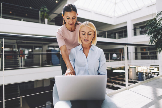 Two smiling diverse businesswomen sit in a large bright office space. Together they look at a laptop and talk about some work.