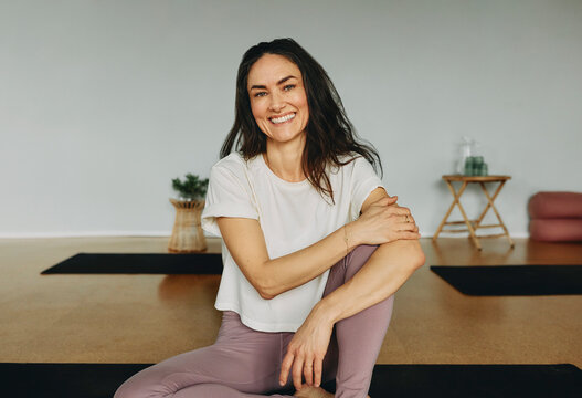 Fit young female yoga instructor in sportswear smiling while sitting on an exercise mat in her studio before a class