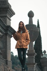 Young woman in a brown coat with fur trim walks along a stone wall in an urban setting.0