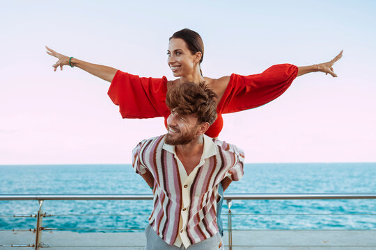 Carefree couple having fun with a piggyback ride by the sea, woman spreading her arms like a bird, enjoying freedom and happiness. - Powered by Adobe