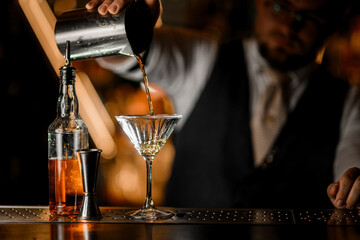 Geyser bottle, a jigger and a tall stem glass are on the bar counter, into which a bartender pours a cocktail