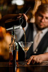 Male bartender pours a cocktail prepared in a mixing glass into a high-stemmed glass
