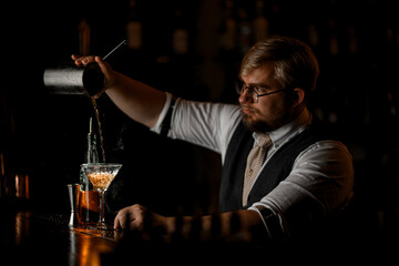 Serious male bartender pours a cocktail prepared in a mixing glass through a strainer into a tall stemmed glass