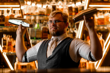 Smiling male bartender holds two shakers above his shoulders like dumbbells, looking at camera