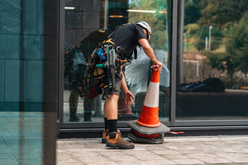 Worker in safety gear places traffic cone near glass building at urban construction site while...