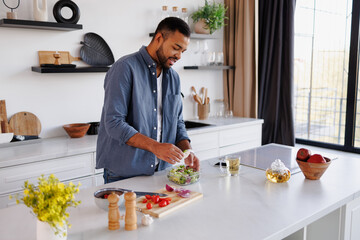 Positive african american man preparing fresh salad near tea in kitchen at home